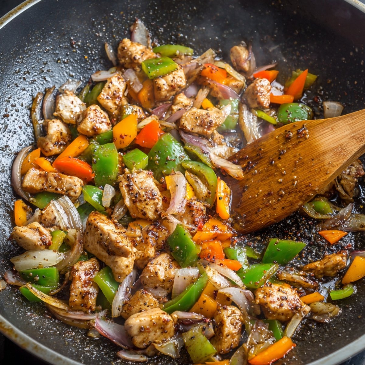 A stir-fried dish in a wok, containing chicken pieces, green and red bell peppers, and onions. The ingredients are being stirred together, with a slight smokiness from the stir-frying process.