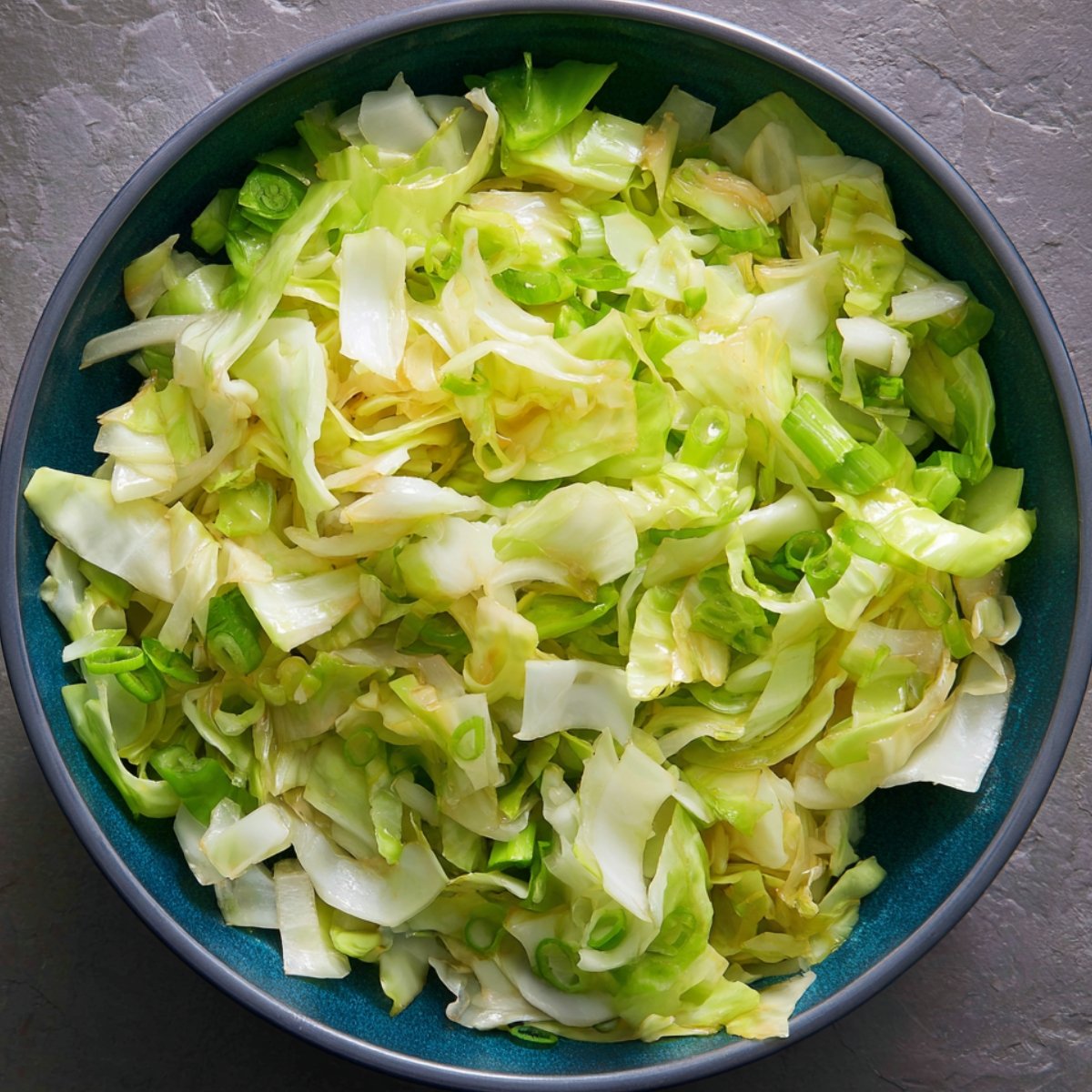 "A close-up image of finely shredded cabbage and green onions in a blue bowl. The cabbage appears to be fresh and crisp, with light green and white hues, while the green onions are scattered throughout."