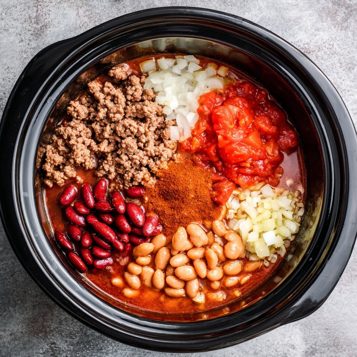 "An overhead view of a crockpot with various ingredients for chili, including ground beef, red kidney beans, white beans, diced tomatoes, chopped onions, and chili powder."