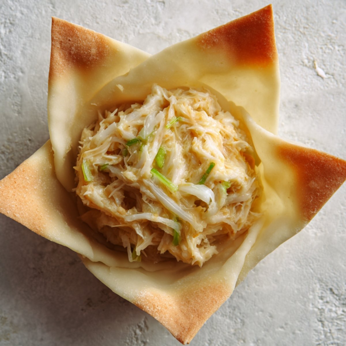 A close-up of a single Rangoon crab bomb sitting on a surface with a filling of shredded crab, cabbage, and green onions inside a crispy, golden wrapper.