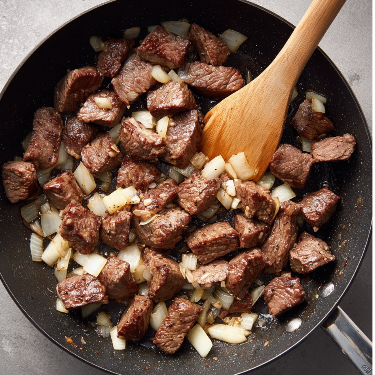 A pan with seared pieces of beef and onions being cooked together, with a wooden spoon stirring the ingredients.