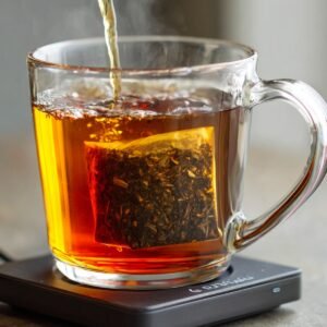 Hot water being poured into a clear glass cup containing a tea bag, with the tea steeping and turning the water amber.