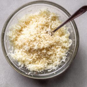 Glass bowl filled with grated Parmesan cheese, seasoned with black pepper, with a spoon inside.