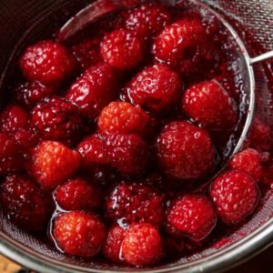 A close-up of juicy raspberries in a strainer, glistening with juice, prepared for making raspberry tea or syrup.