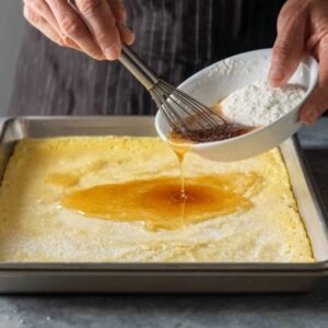 Hands holding a whisk and bowl, pouring vanilla mixture over a baked sheet pan pancake.