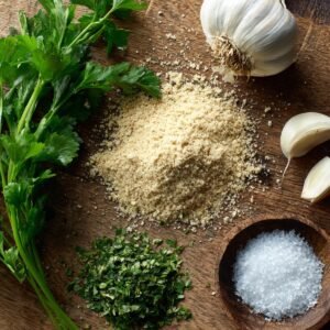 Fresh parsley, garlic cloves, a bulb of garlic, coarse salt, chopped herbs, and grated Parmesan cheese arranged on a rustic wooden board.