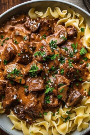 "A serving of Beef Stroganoff with tender beef chunks in a creamy sauce, served over egg noodles and garnished with parsley."