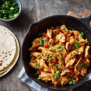 A skillet filled with seasoned grilled chicken strips, diced tomatoes, and cilantro, served alongside warm flour tortillas ready for assembling a crunch wrap.