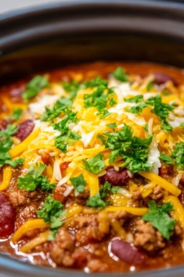 A crockpot Chili filled with a hearty chili made of ground beef, kidney beans, diced tomatoes, and topped with shredded cheddar cheese and chopped cilantro as garnish.
