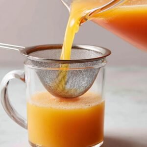 Freshly blended peach lemonade being poured through a fine mesh strainer into a glass mug to remove pulp.