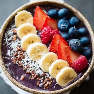 A finished acai bowl recipe topped with banana slices, strawberries, raspberries, blueberries, shredded coconut, and crunchy granola.
