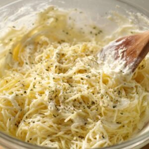 "Spaghetti mixed with cream cheese and herbs, being stirred with a wooden spoon in a glass bowl."