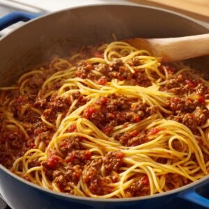 "A close-up of cooked spaghetti mixed with a rich meat sauce in a large pan, with steam rising."
