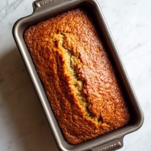 A freshly baked golden-brown loaf of banana bread in a rectangular metal pan, with a split down the center showing its moist texture.
