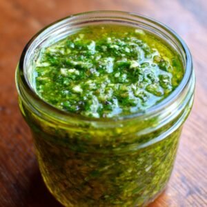 A glass jar filled with basil pesto, its surface glistening with olive oil, showing a chunky texture of herbs, nuts, and cheese.