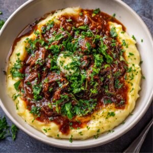 A close-up of a bowl of mashed potatoes topped with rich, savory beef stew, garnished with fresh chopped parsley.