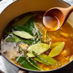 A pot filled with simmering broth, bay leaves, and thyme, with a wooden spoon pouring liquid into it.