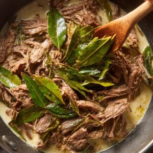 A close-up of cooked beef stew with shredded beef and bay leaves in a creamy sauce.