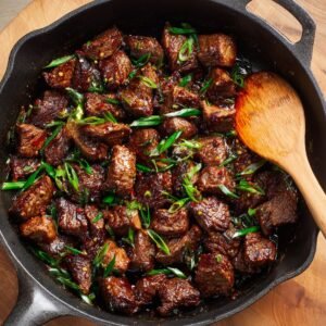"Tender beef cubes in a rich sauce with green onions on top, cooking in a cast iron skillet."