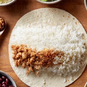 Tortilla topped with a line of fluffy white rice and seasoned shredded chicken, showing the first step of burrito assembly.