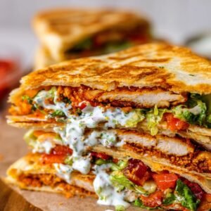 A close-up of a crispy golden-brown chicken crunch wrap, sliced in half to reveal layers of fried chicken, fresh lettuce, diced tomatoes, melted cheese, and creamy sauce wrapped in a toasted tortilla.