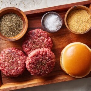 "Three seasoned raw burger patties on a wooden tray with a sandwich bun and small bowls of black pepper, salt, and mustard seeds."