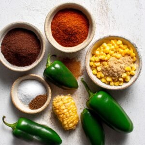 A collection of chili ingredients and spices, including green chili peppers, paprika, corn, cumin, and salt, arranged on a clean surface.