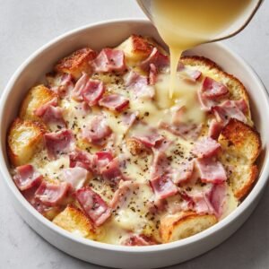 Unbaked eggs benedict casserole with chunks of bread, diced ham, melted cheese, and egg mixture being poured in.