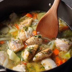 A simmering pot of fish soup with chunks of white fish, carrots, leeks, and herbs in a clear broth.
