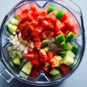 "Fresh chopped vegetables including tomatoes, cucumbers, green bell peppers, onions, garlic, and spices inside a blender jar before blending."