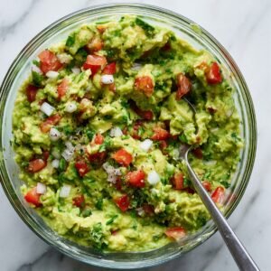 A glass bowl filled with chunky guacamole mixed with diced tomatoes, onions, cilantro, and black pepper.