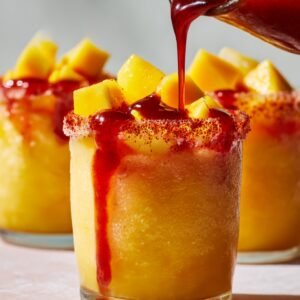 Mangonada served in a glass rimmed with Tajín, filled with frozen mango slush, topped with mango chunks, while chamoy sauce is being drizzled over the top.
