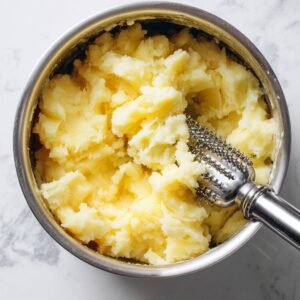 A close-up of mashed potatoes being mashed with a potato masher in a stainless steel bowl.