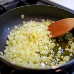 A pan of diced onions being sautéed in olive oil with a wooden spoon.
