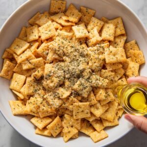 A white bowl filled with square cheese crackers topped with parmesan cheese, herbs, and spices, with a hand pouring olive oil over them.