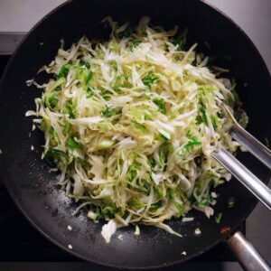 Stir-fried cabbage with some chopped green onions in a black pan with tongs.