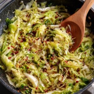 Stir-fried cabbage with red pepper flakes and a wooden spoon stirring it in a black pan.