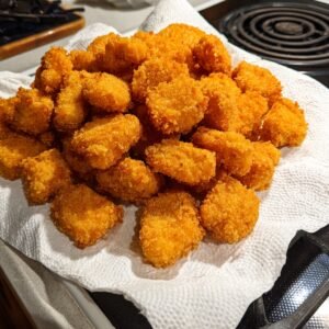 A pile of golden-brown fried chicken nuggets resting on a paper towel to absorb excess oil, placed on a kitchen counter near a stovetop.