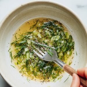 Small bowl of homemade cucumber salad dressing with vinegar, dill, chopped onion, and black pepper being mixed with a fork.