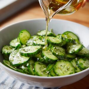 Cucumber slices in a white bowl with a light dressing being poured over them, garnished with herbs for a fresh, tangy salad.