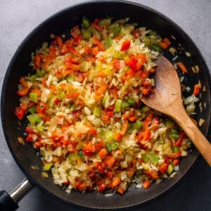 A pan of sautéed onions, red bell peppers, and green bell peppers, with a wooden spoon stirring the colorful mixture.