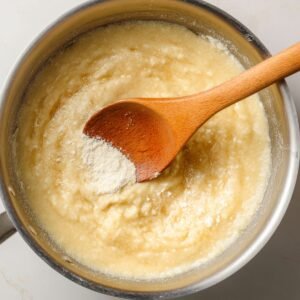 Thick churro dough cooking in a saucepan, flour being mixed into the butter base with a wooden spoon.