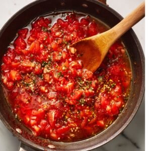 Pan of diced tomatoes simmering with spices and seasonings, stirred with a wooden spoon.