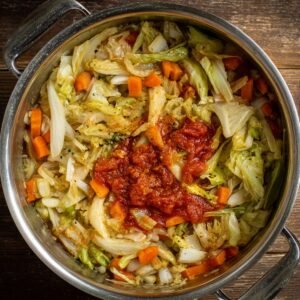 A pot of cabbage soup being prepared, with chopped cabbage and carrots simmering, topped with tomato sauce and seasonings before stirring.
