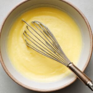 A bowl of creamy yellow mixture, possibly custard or batter, being whisked with a metal whisk in a ceramic bowl on a light surface.