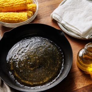 A cast iron skillet with oil heating, next to fresh corn on the cob, a glass jar of oil, and a stack of paper towels on a plate.