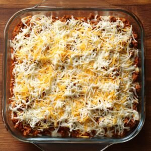 Glass casserole dish layered with seasoned beef, sour cream mixture, and shredded cheese before baking.