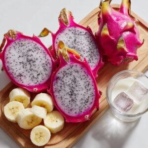 "Dragon fruit halves, banana slices, and a glass of milk with ice cubes arranged on a wooden board for smoothie preparation."