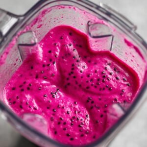 "Blender container with thick, creamy dragon fruit smoothie showing a vibrant pink color with black seeds."