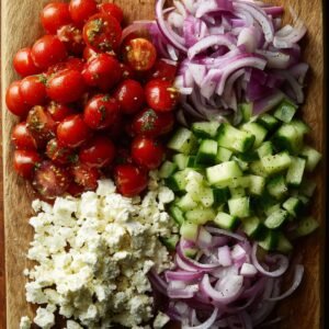 Close-up of chopped cucumbers, cherry tomatoes, sliced red onion, and crumbled feta cheese neatly arranged on a cutting board, seasoned with olive oil and herbs.